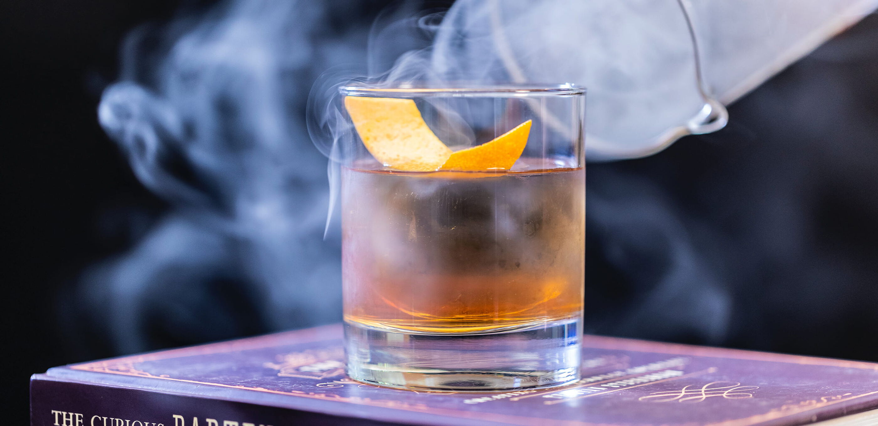 A smoking cloche is taken off an old fashioned cocktail, filling the air with aromatic smoke